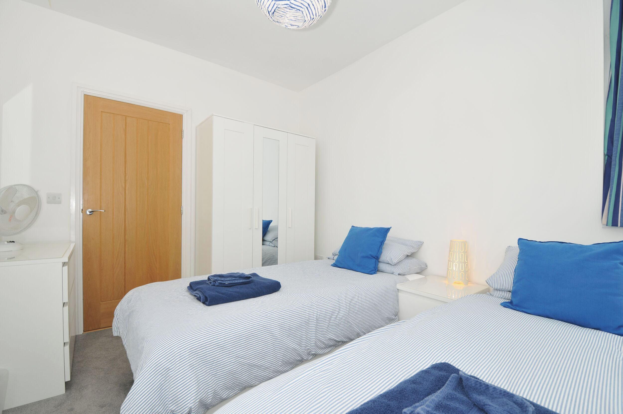 Une chambre avec deux lits, une porte en bois, une armoire blanche, une table de chevet avec une lampe et un ventilateur posé sur une commode.