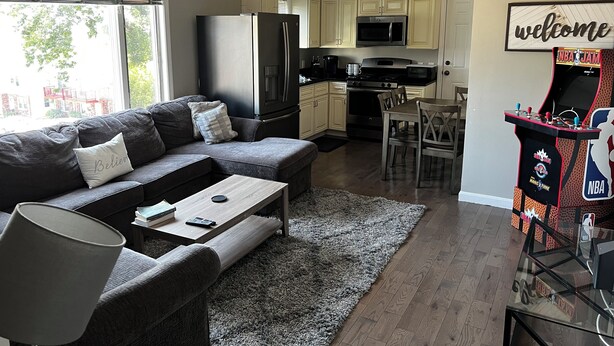 A modern living room with a sofa, coffee table, and a small table with books and a lamp. There is a kitchen area with cabinets, a refrigerator, and a microwave. A welcome sign is on the wall, and a basketball arcade game is visible. - Charming 2-bedroom condo in delightful Chelsea with WiFi, AC (Chelsea)