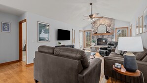 A living room with a stone fireplace, leather sofas, a television, and a ceiling fan. - Oak Haven Retreat, Oak Haven Retreat  - Charming Green Lake Property (Princeton)