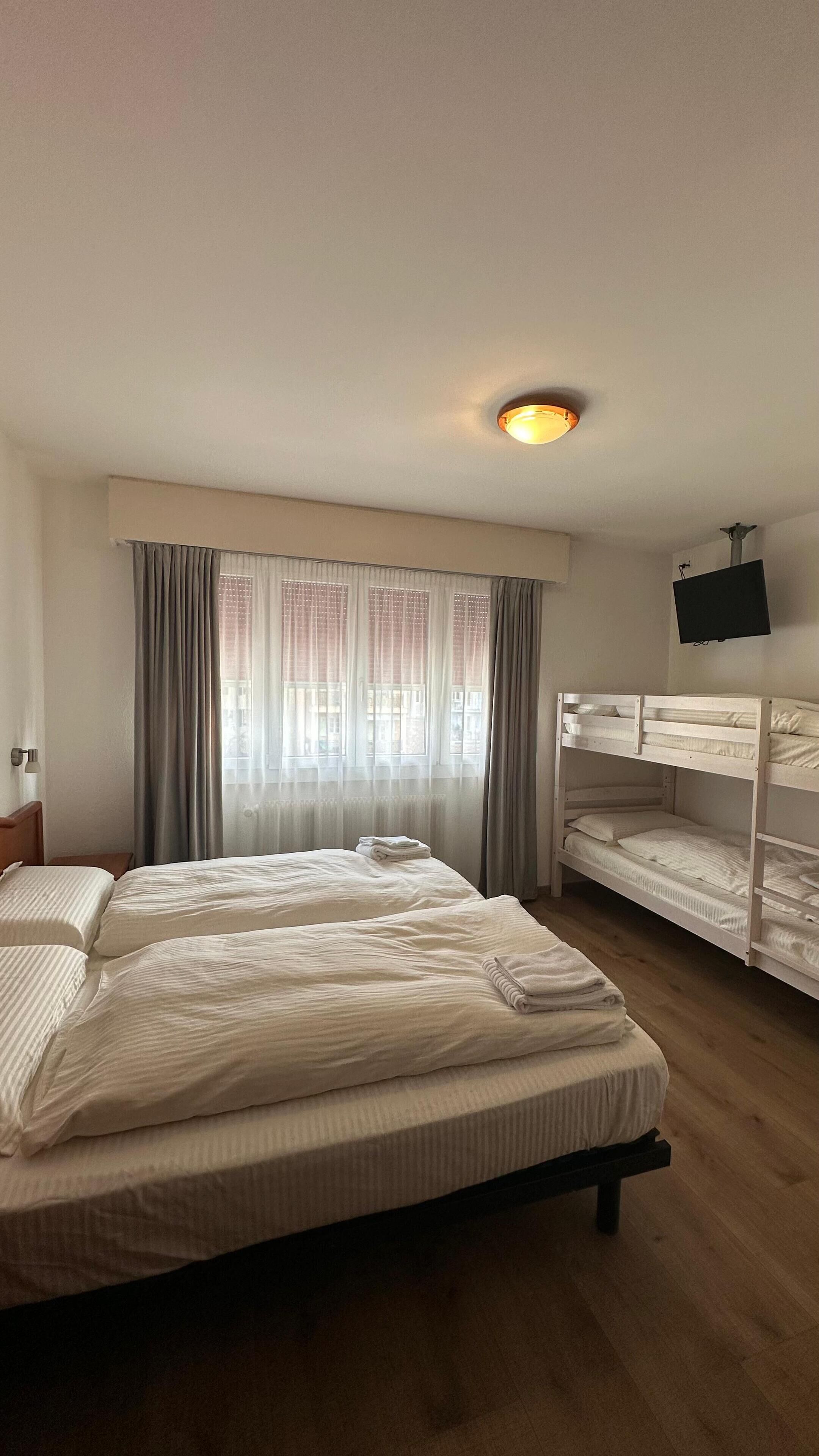 Habitación de hotel con una cama grande, una cama litera, un televisor de pantalla plana montado en la pared y una ventana con cortinas.