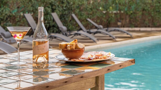 A table with a bottle of La Cote, a martini glass, and a plate of food by a poolside.