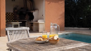 A wooden table with a plate of croissants and a pitcher of orange juice, a glass of orange juice, and a bowl of bread by a poolside.