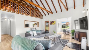 Un ampio soggiorno con soffitto alto e travi a vista in legno, un divano grigio, un tavolino da caffè e un'area pranzo con una TV.