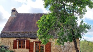 A stone house with a sloped roof, wooden shutters, and a wooden door. There is a tree in front of the house and a picnic table with chairs. - Typical Périgord Noir house with private pool (Saint-Geniès)