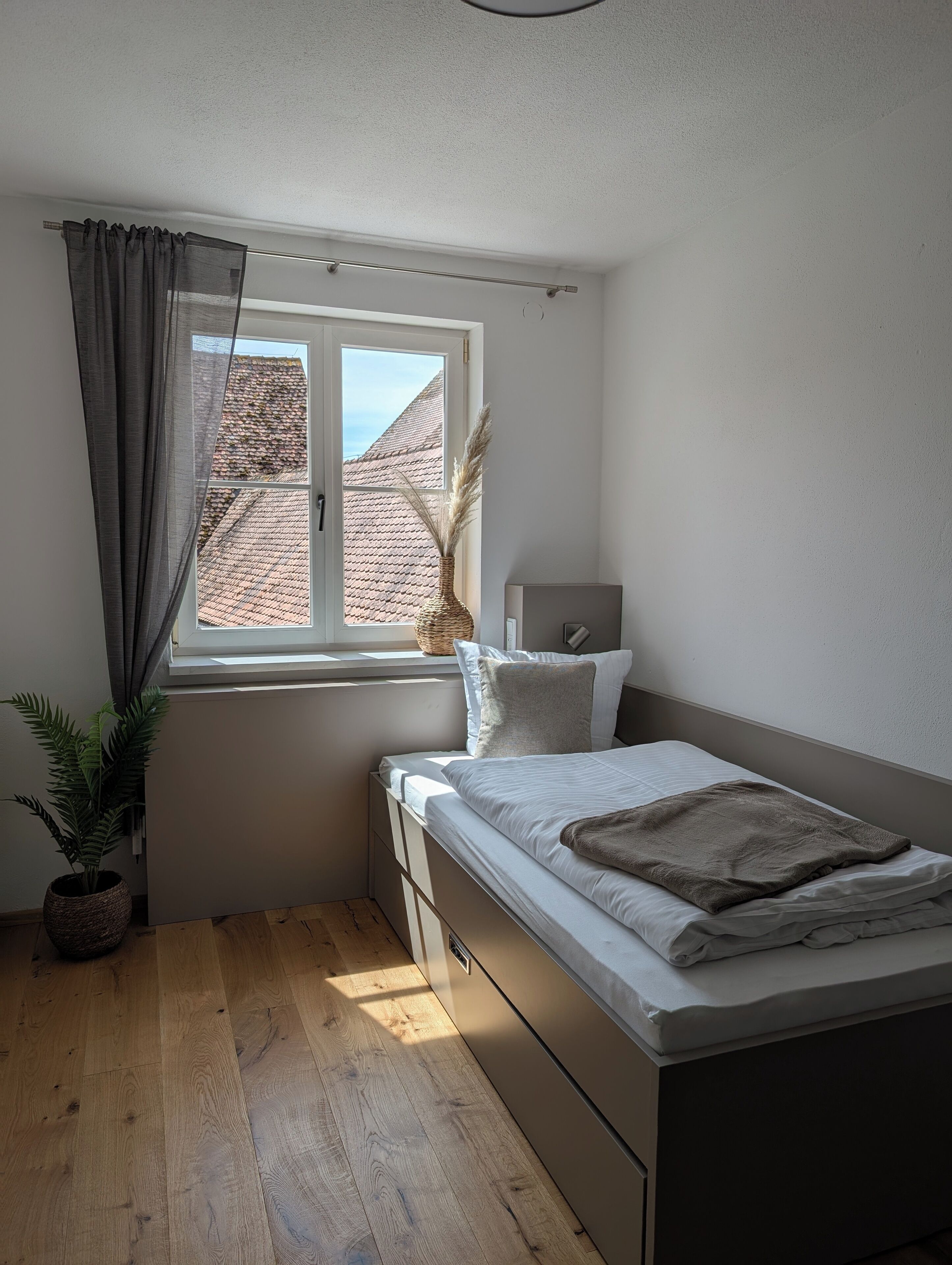 Een slaapkamer met een bed, een raam met gordijnen, een plant en uitzicht op een gebouw.