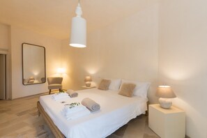 Una cama bien hecha con toallas enrolladas, una lámpara de noche y una silla en una habitación luminosa con un espejo grande en la pared.