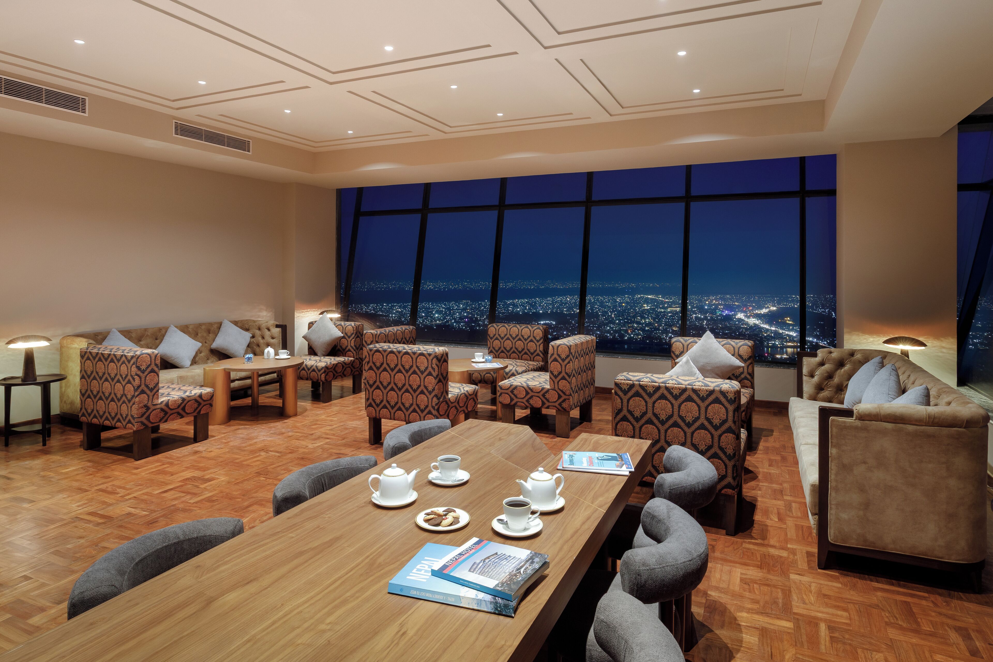 A modern hotel lobby with a large wooden table set for tea, patterned sofas, and a panoramic city view.