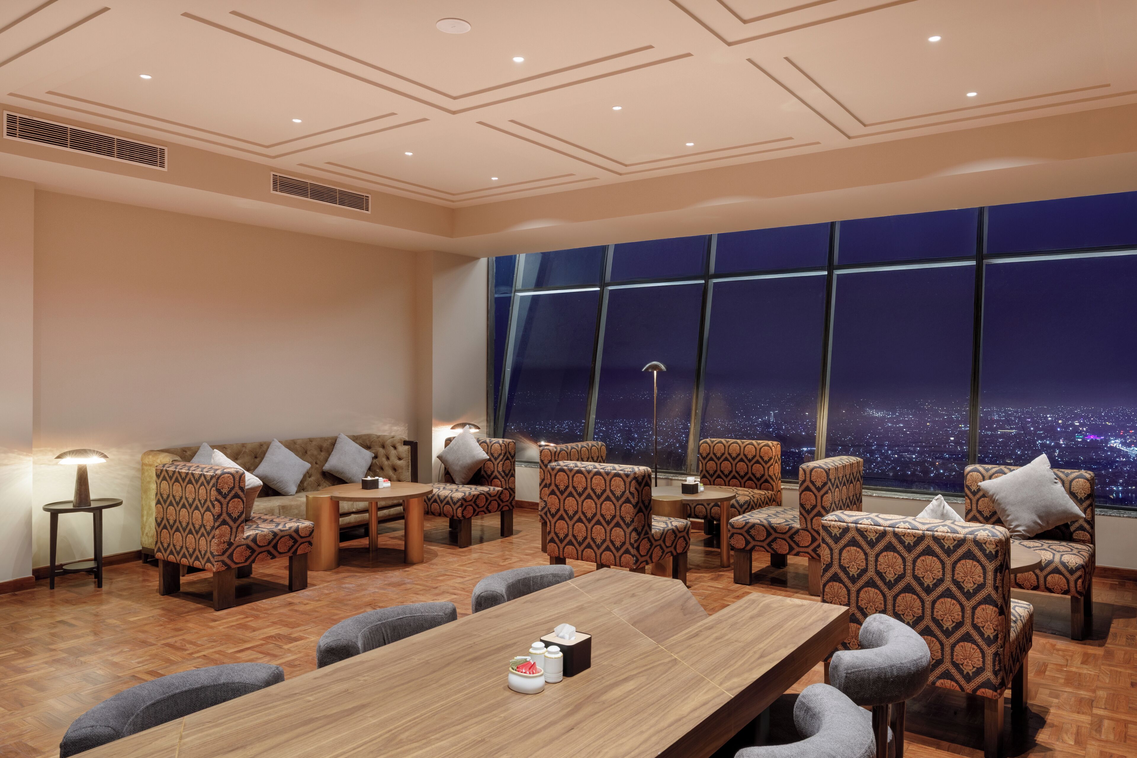 A hotel room with a large window offering a city view at night, a sofa, multiple patterned chairs, a wooden table, and a lamp.