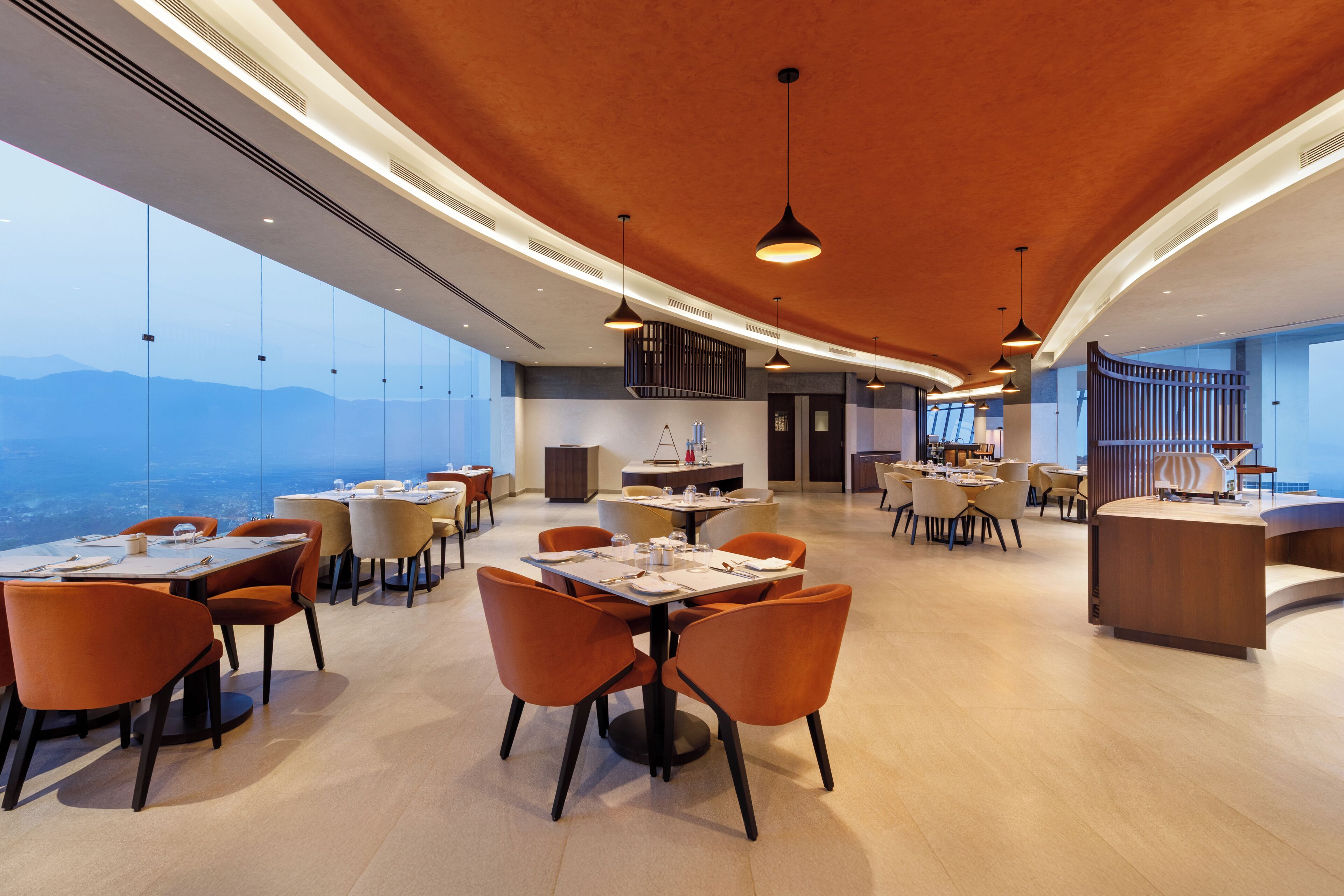 A spacious dining area with high tables and orange chairs, large windows offering a view of the mountains, and a modern ceiling design.