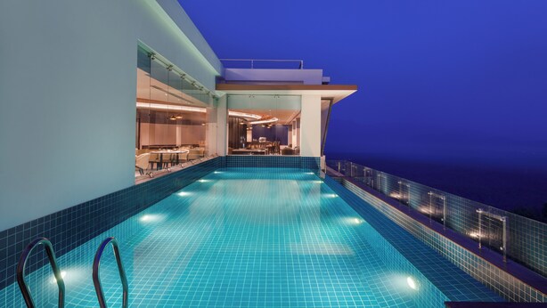 Una piscina infinita moderna con baldosas azules cristalinas, barandilla de vidrio y vista al océano al atardecer.