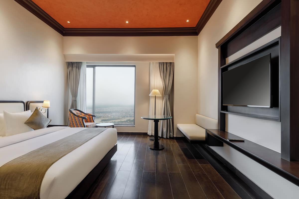 A hotel room with a large bed, a TV, a seating area, and a view of the city.