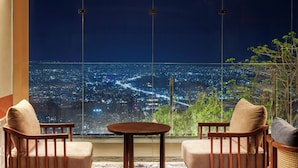 Una habitación con vista a la ciudad, dos sillas de madera, una mesita redonda y un ventanal grande.