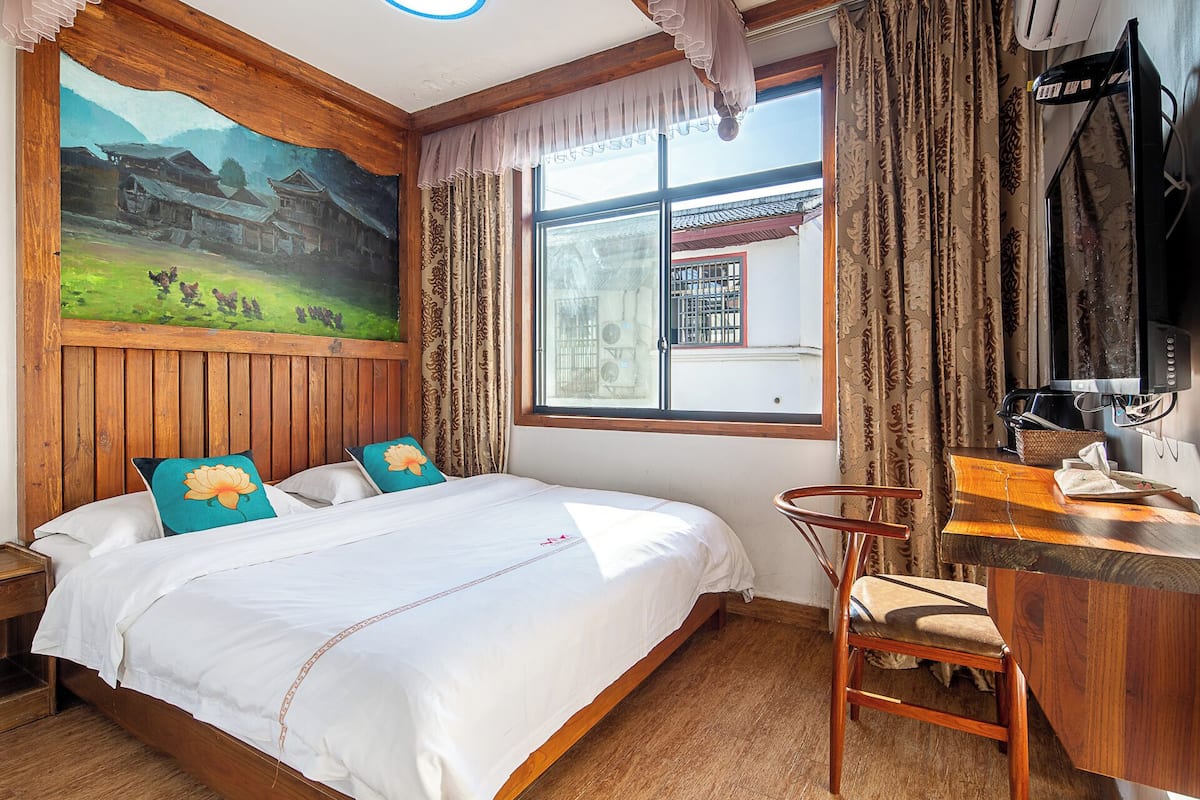 A bedroom with a large bed, wooden headboard, a painting of a landscape, a window with curtains, and a desk with a chair.