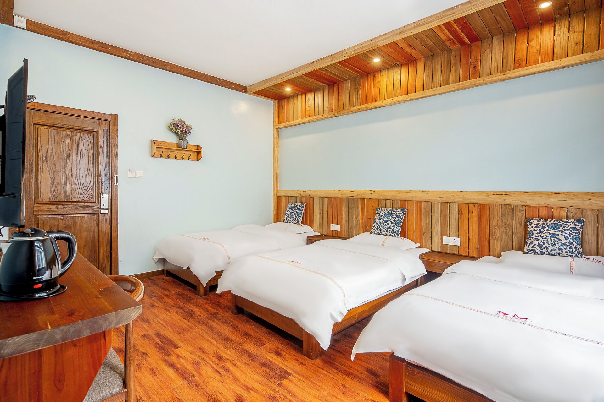Ein Hotelzimmer mit drei Betten, Holzvertäfelung, einem Holztisch mit Wasserkocher und einem Wandregal mit Haken.
