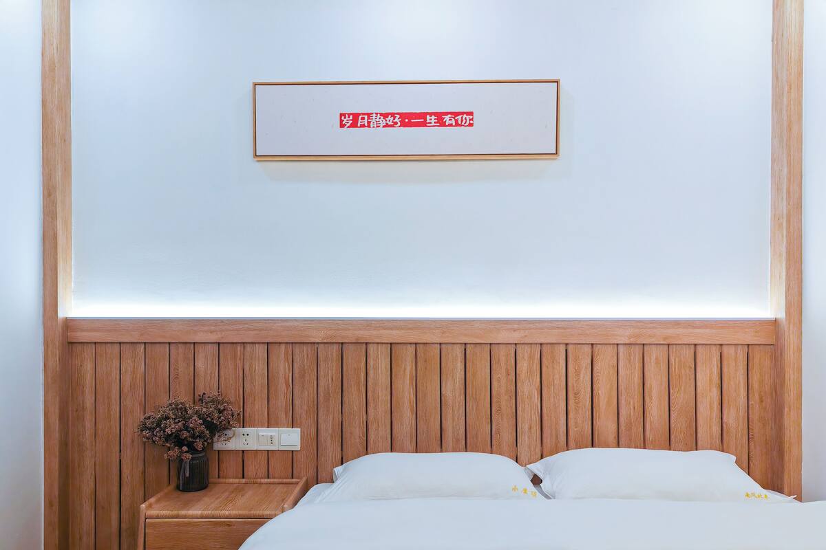 A neatly arranged bedroom with a wooden headboard, a bedside table with a vase of dried flowers, and a sign above the bed.