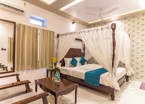A bedroom with a four-poster bed, a bedside table with a vase and bottles, a wooden bench, and a ceiling fan. - Lake N Hill View (Udaipur)