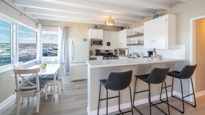 A modern kitchen with white cabinetry, a central island with bar stools, a dining area with a table and chairs, and a view of the city through large windows. - Stylish Newport Condos w/ Dock Access, Walk to Balboa Fun Zone & Beach (Newport Beach)