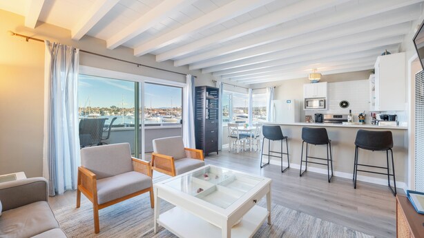 A modern living room with a glass coffee table, a sofa, and a dining area with a view of the marina. - Stylish Newport Condos w/ Dock Access, Walk to Balboa Fun Zone & Beach (Newport Beach)