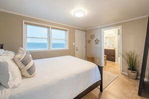A bedroom with a large bed, a view of the sea through a window, and a bathroom with a vanity and mirror.