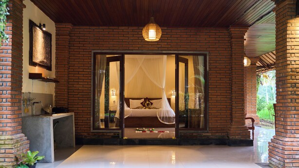 A spacious room with a large bed, a small table, and a chair. There is a window with a view of greenery outside. - Hening Suite Ubud (Tegallalang)