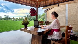 A person seated at a wooden table with a laptop, a potted plant, and a red flower in a thatched-roof room with a view of a green field. - Hening Suite Ubud (Tegallalang)