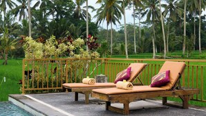 A poolside area with two lounge chairs and a small table, surrounded by a wooden fence and palm trees. - Hening Suite Ubud (Tegallalang)