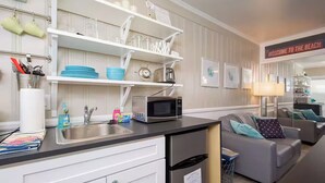 A compact kitchen with a microwave, sink, and storage shelves. There is a couch and a sign that reads "Welcome to the Beach" in the background.