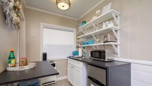 A compact kitchen with white cabinets, a black countertop, and a microwave. There is a small window with blinds, a blue chair, and a bottle of soda on the counter.