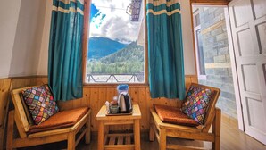 A cozy room with two wooden chairs, a small table with a kettle, cups, and a tray, and a window with curtains offering a mountain view. - Singh Saab Rooms & Cafe (Manali)