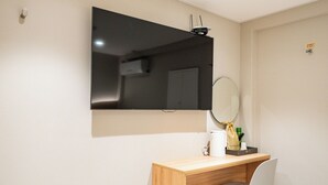 A small, modern room with a wooden desk, a white chair, a mirror, and a flat-screen TV mounted on the wall. - Cream Hotel (Guri)