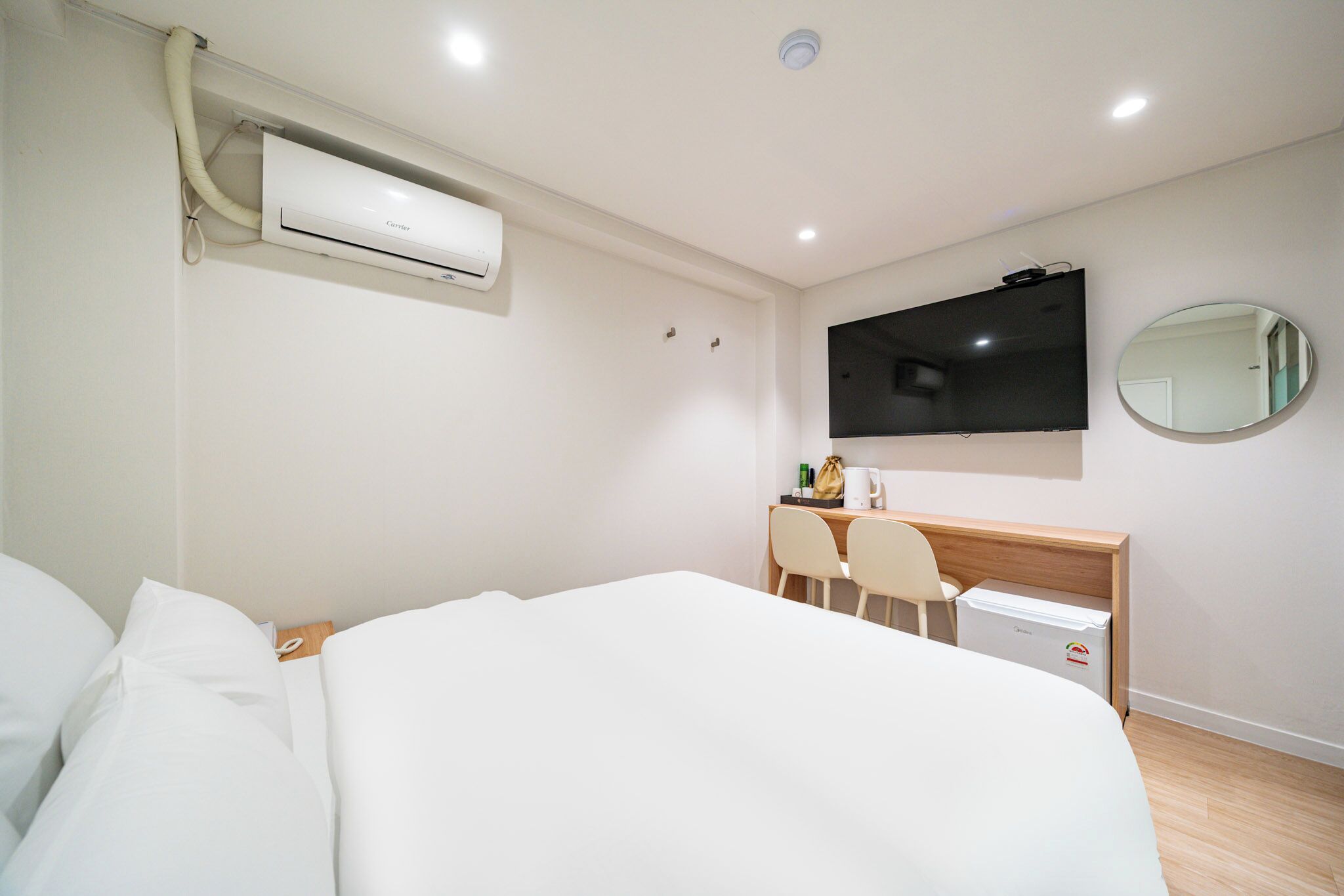 A hotel room with a bed, a desk with a chair, a television, a mirror, and an air conditioning unit.