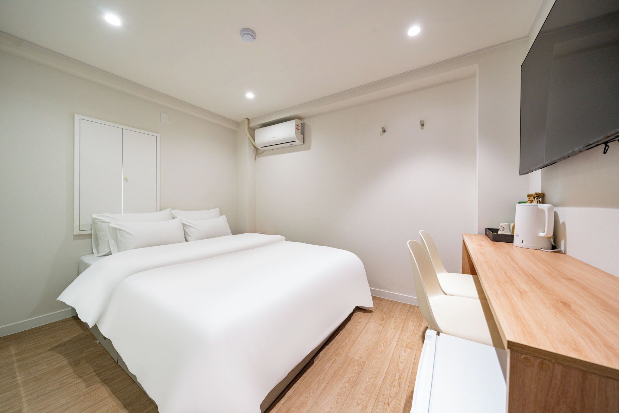 A hotel room with a bed, a desk, a chair, a flat-screen TV, and an air conditioning unit.