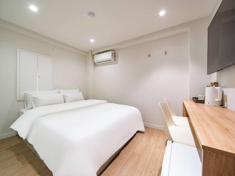 A hotel room with a bed, a desk, a chair, a flat-screen TV, and an air conditioning unit.
