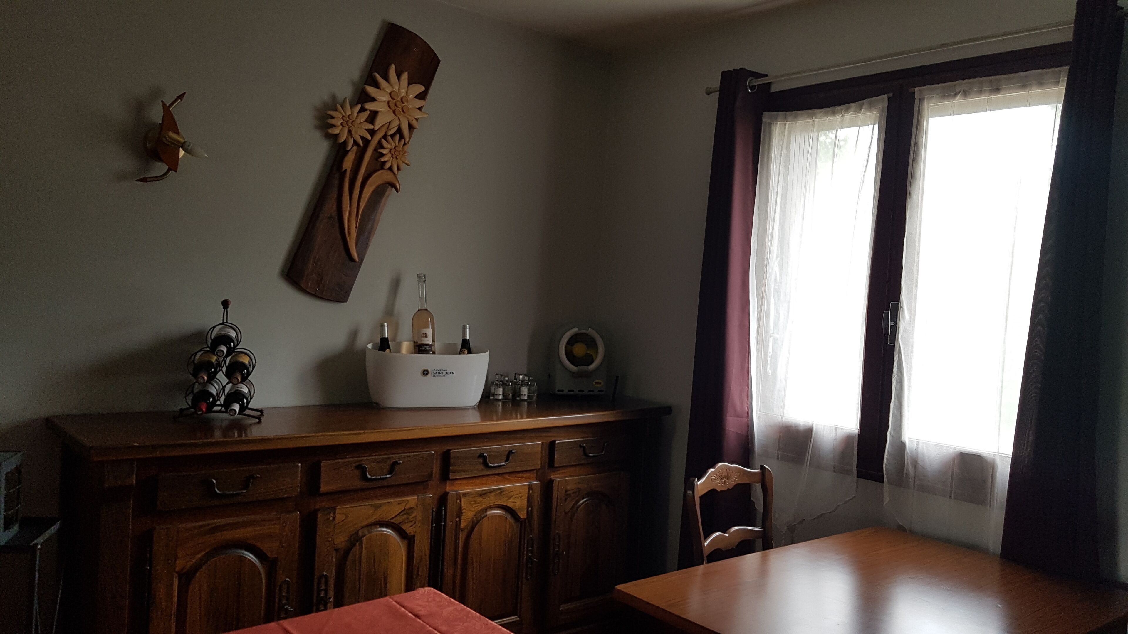 Een eetgedeelte met een houten tafel en stoelen, een wijnrek, een decoratief wandkunstwerk en een raam met gordijnen.