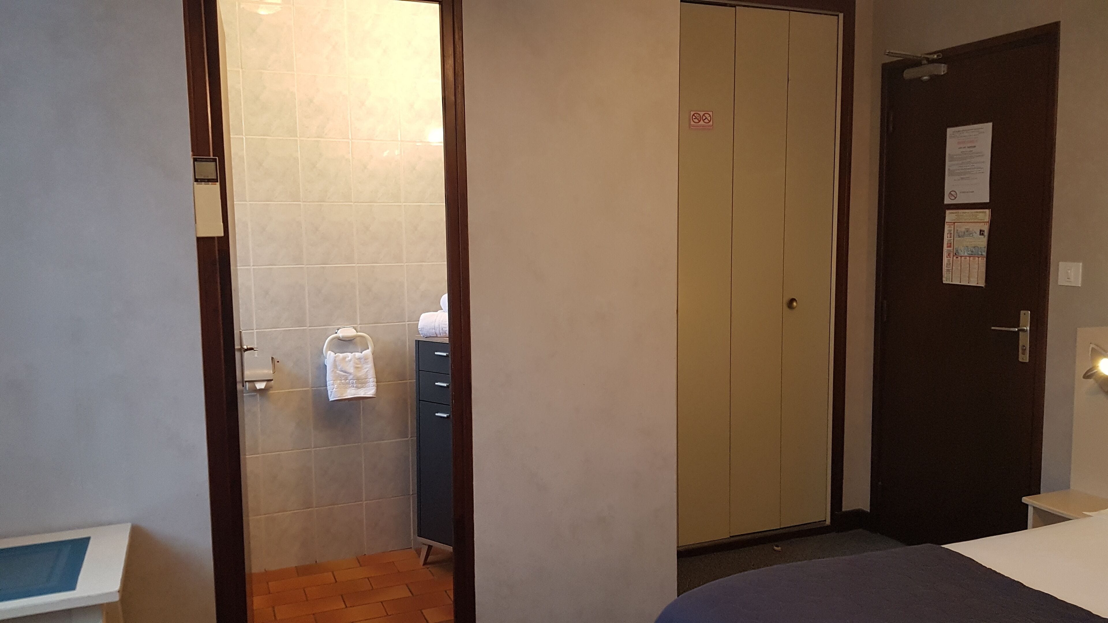 Une chambre d’hôtel avec un plancher en bois, un lit, un bureau, une armoire et une salle de bain dont un mur est recouvert de carreaux et sur lequel est accrochée une serviette.