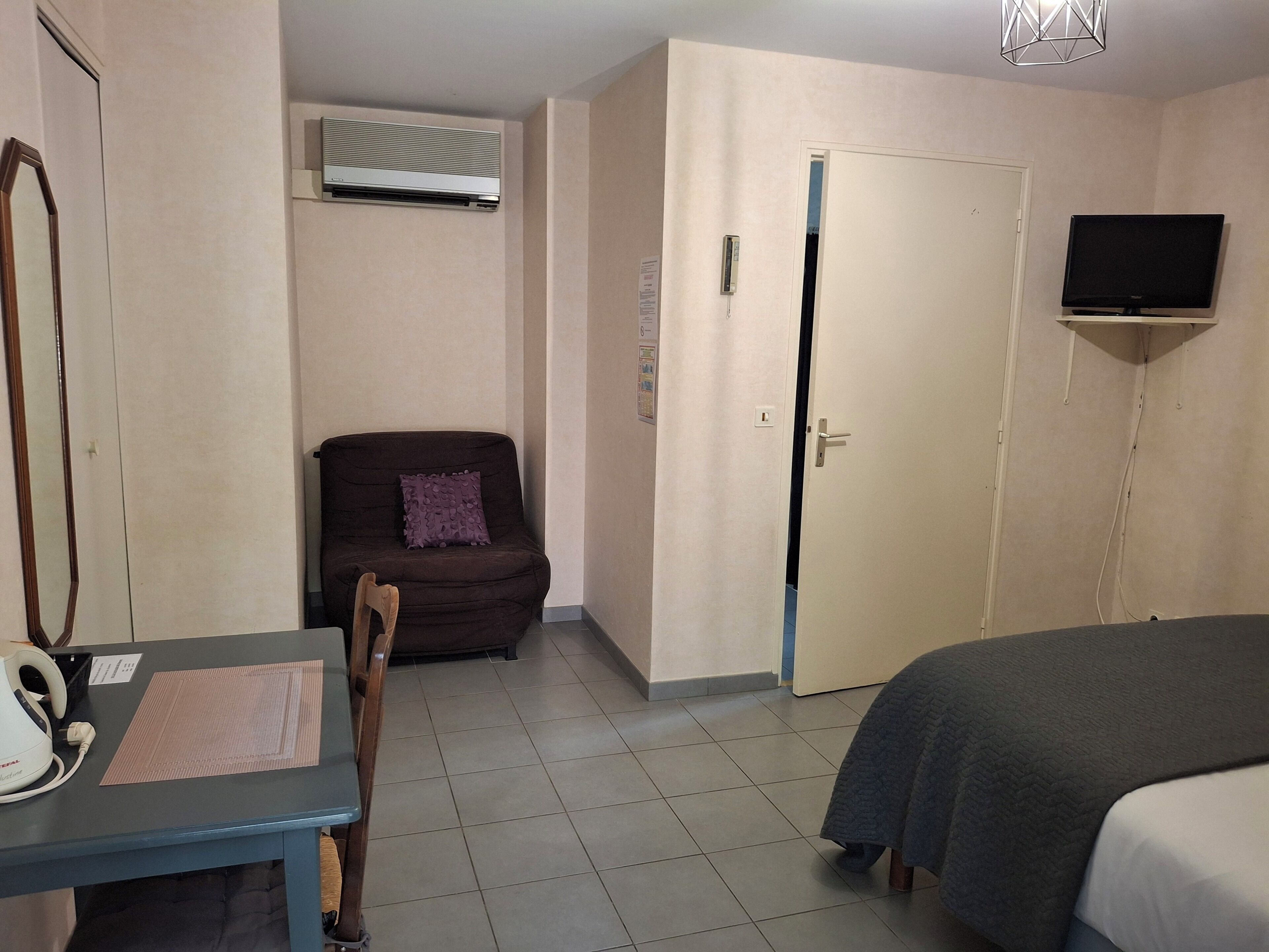 Habitación de hotel con cama, escritorio con silla, sofá, unidad de aire acondicionado, televisor montado en la pared y una puerta.