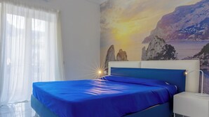 A bedroom with a bed, a nightstand, a wall mural of a coastal scene, and a window with sheer curtains. - CapriGetaway - Domus Le Botteghe (Capri)