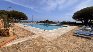 Una piscina rectangular con solárium de piedra, rodeada de tumbonas y sombrillas, con vistas al mar.
