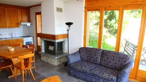 A cozy living area with a fireplace, a sofa, a dining table with chairs, and a large window offering a view of greenery. - Alluring apartment in Les Agettes (Les Agettes)