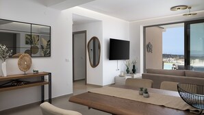 A modern living room with a wooden dining table, beige sofas, a TV mounted on the wall, and a large window offering a view of the outdoors.
