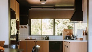 A modern kitchen with wooden cabinets, a dining table with chairs, and a view of greenery outside. - The Perfect Apartment (Sandy Bay)