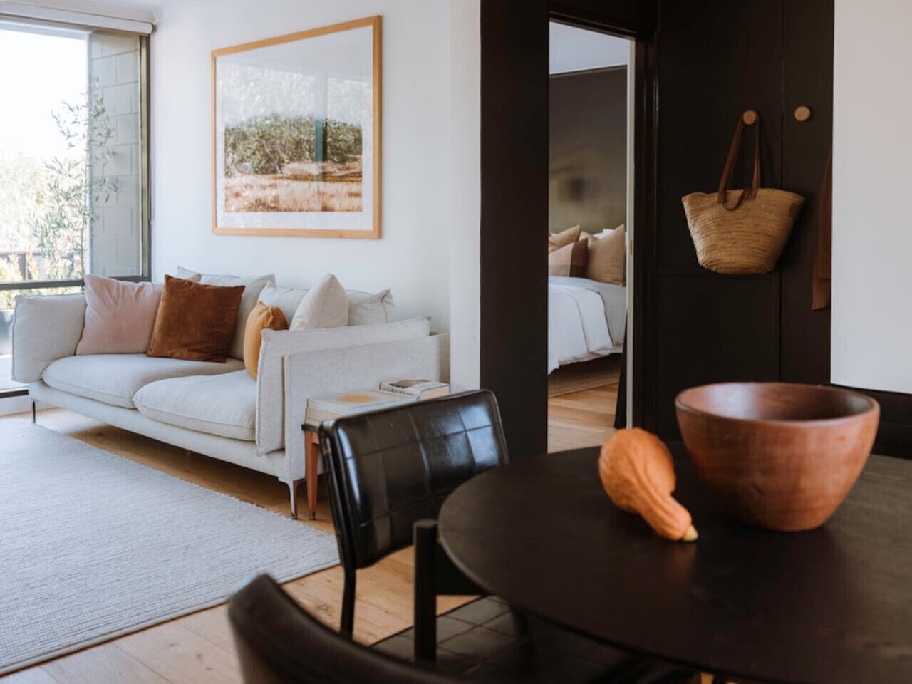 A modern living room with a white sofa, black leather chairs, a wooden table with a bowl and a squash, and a large window with a view of greenery.