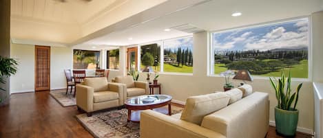 A spacious living room with a large window offering a view of a golf course, a ceiling fan, and a dining area with wooden furniture.