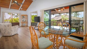 A spacious room with a glass table and wicker chairs, large windows with a view of palm trees, and a ceiling fan.