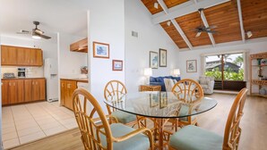 A spacious living area with a glass-top dining table surrounded by wicker chairs, a wooden ceiling, and a view of the outdoors through a large window.