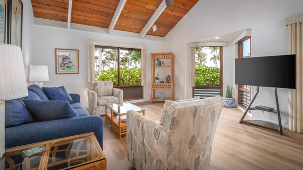 A spacious living room with a high ceiling, wooden beams, a ceiling fan, a large TV, and a view of palm trees through the windows.