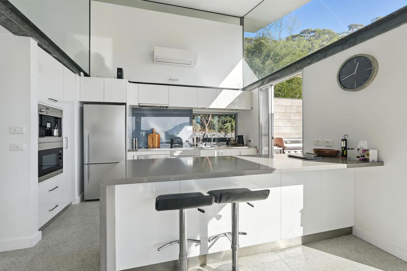 A modern kitchen with white cabinetry, a stainless steel refrigerator, a built-in oven, and a large island with bar stools.