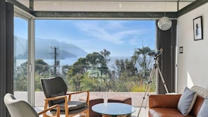 Een kamer met een grote glazen deur die leidt naar een balkon met uitzicht op een meer en bergen. Op het balkon staat een statief met een camera, een bruine leren bank en een ronde glazen tafel.