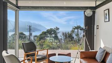 A room with a large glass door leading to a balcony with a view of a lake and mountains. There is a tripod with a camera on the balcony, a brown leather sofa, and a round glass table.