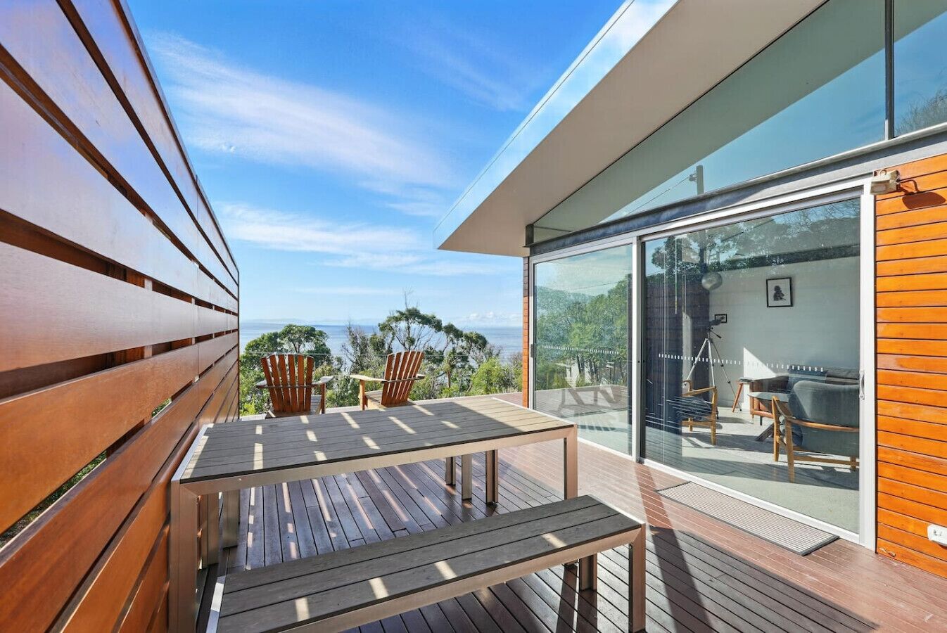 A modern outdoor deck with wooden furniture, a glass door leading to a balcony, and a view of the ocean.